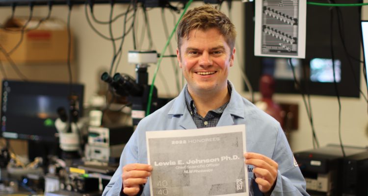 Dr. Lewis Johnson holds up his PSBJ 40 under 40 announcement, background is scientific lab probe station